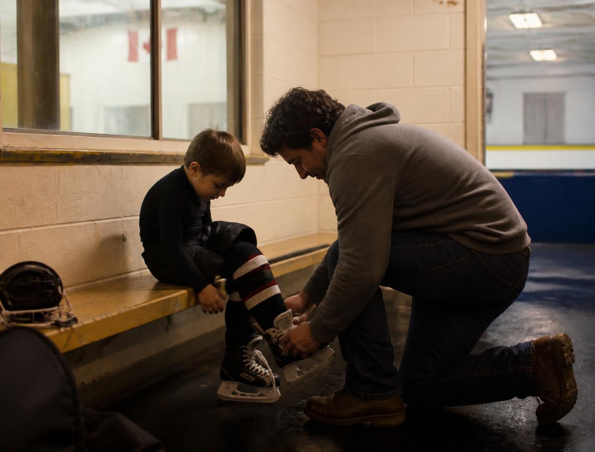 A parent kneeling to help a young child lace up skates on a bench at a community arena
