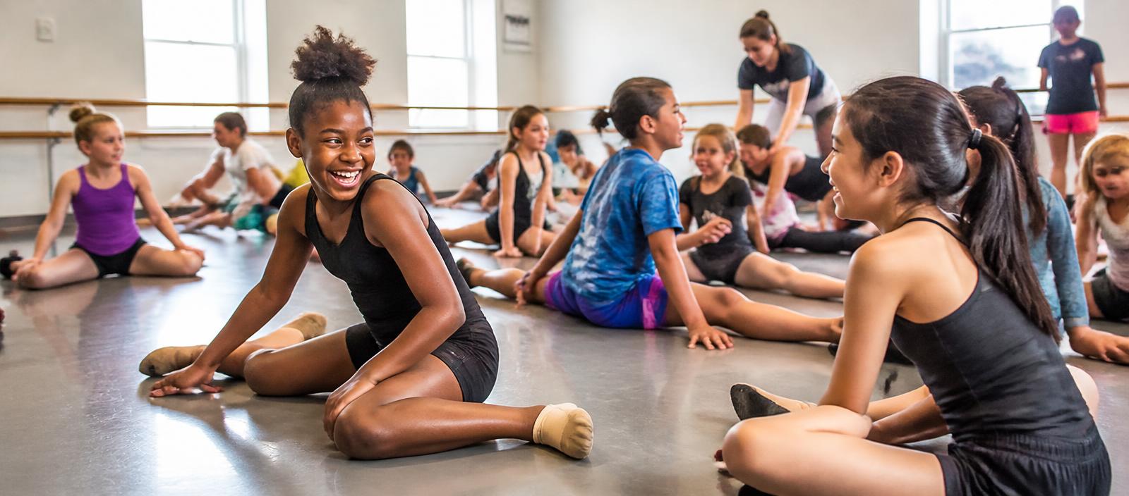 Young dancers and gymnasts sitting on a studio floor stretching and laughing together before class